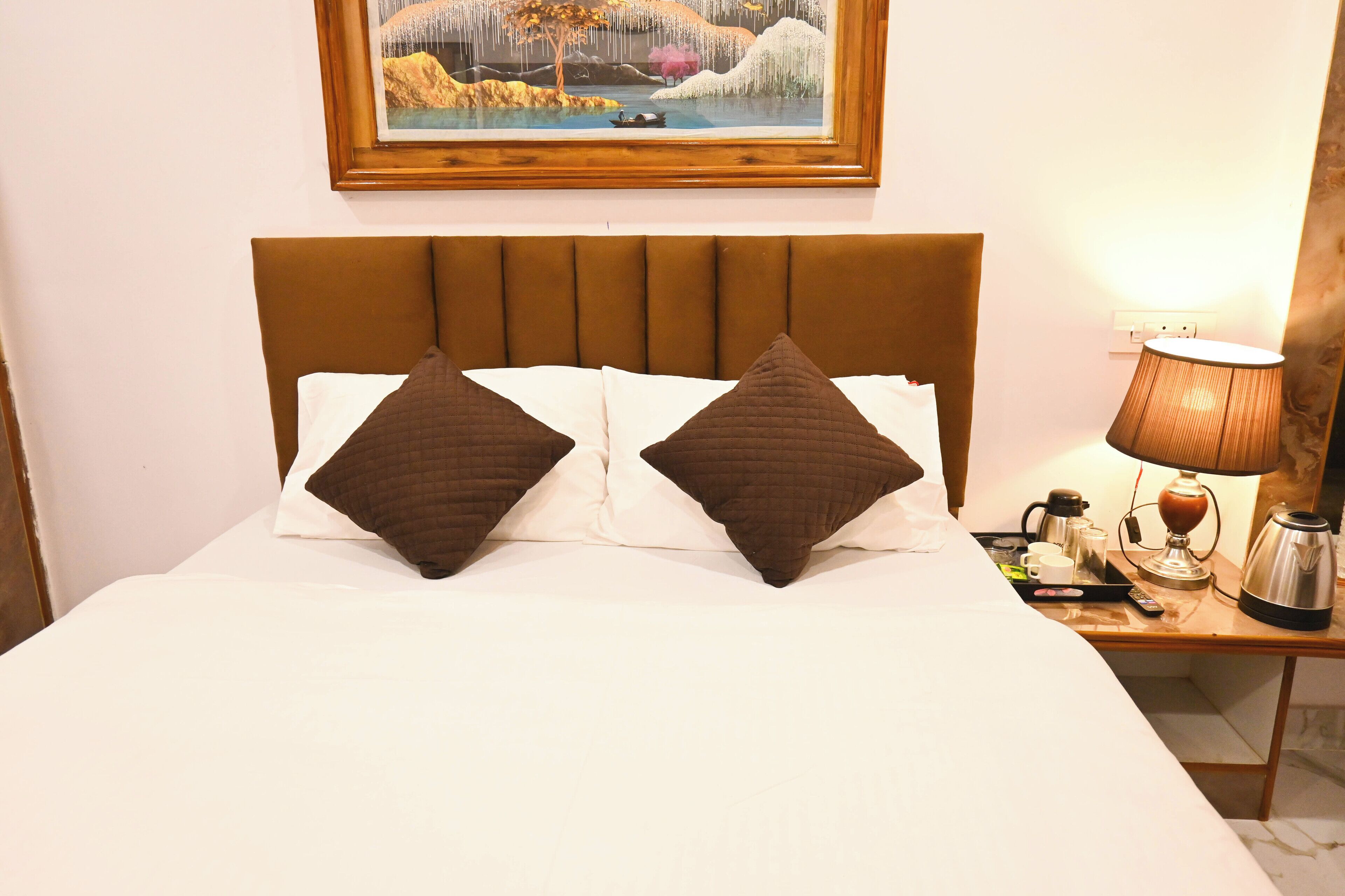 Una cama bien tendida con dos almohadas marrones, una mesita de noche con una lámpara, una cafetera y un cuadro enmarcado sobre la cama.