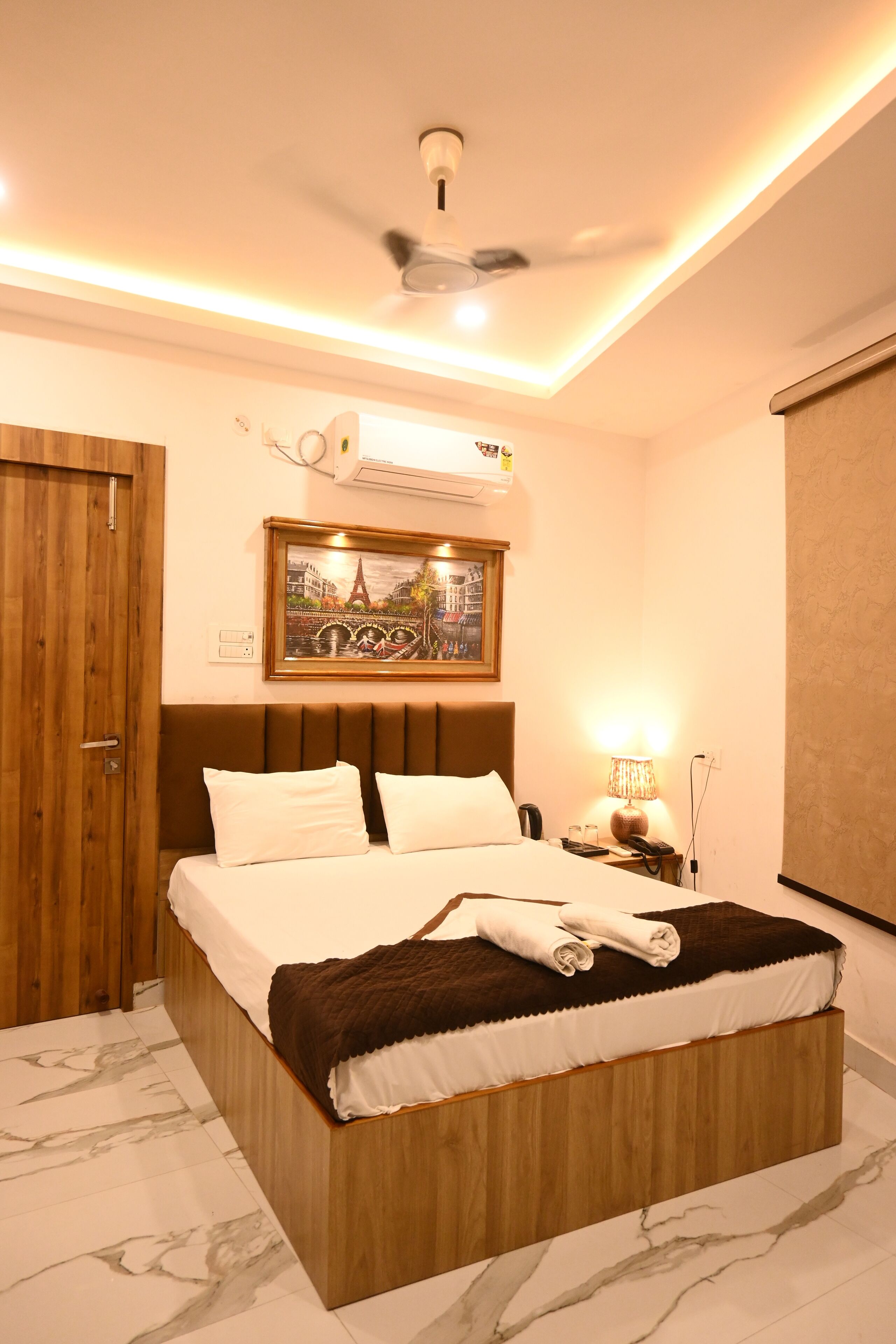 Une chambre à coucher avec un lit, un ventilateur de plafond, un climatiseur fixé au mur, un tableau encadré, une lampe de chevet et une porte en bois.
