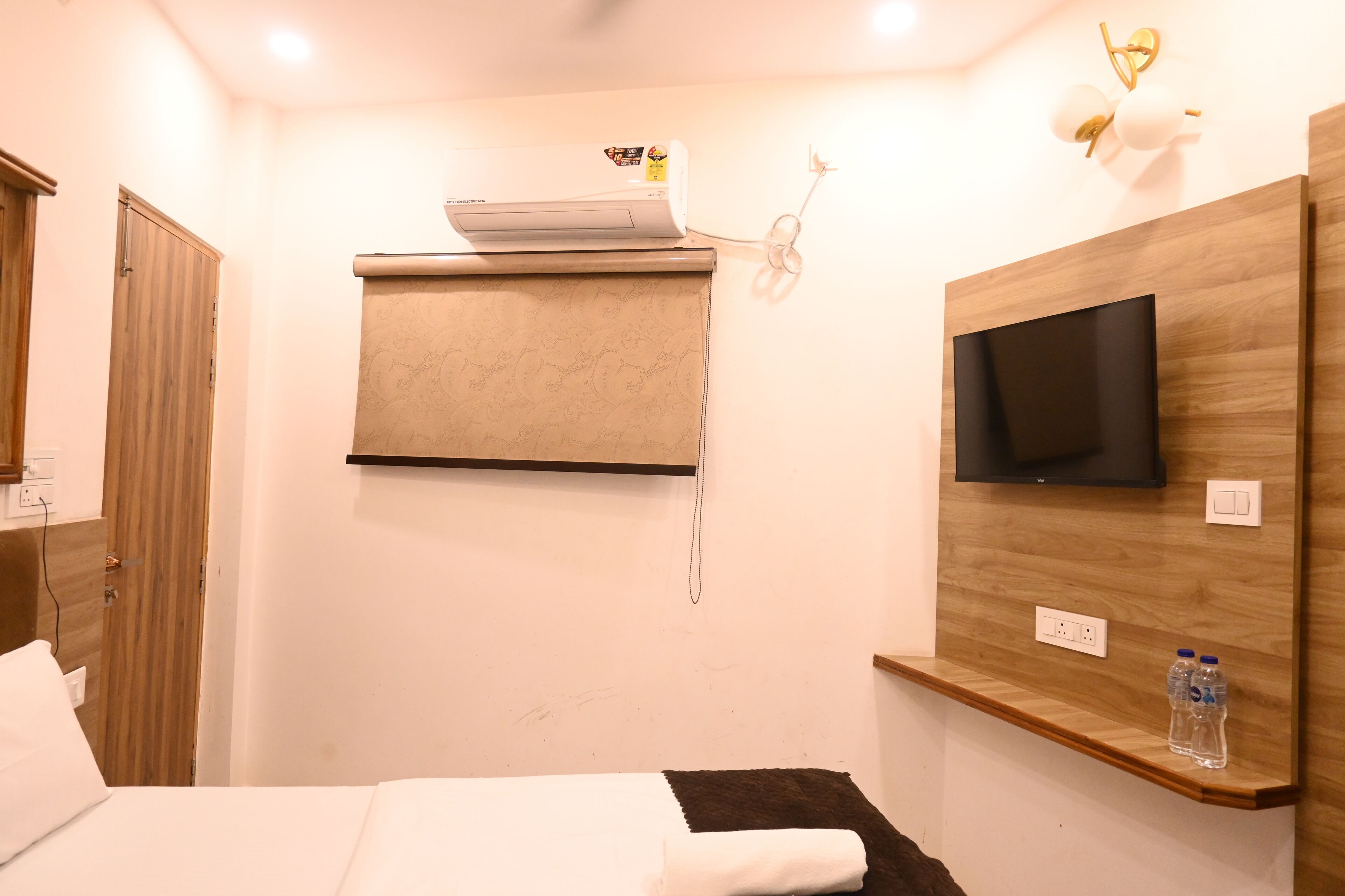 A hotel room with a bed, a mounted air conditioner, a flat-screen TV, and a shelf with water bottles.