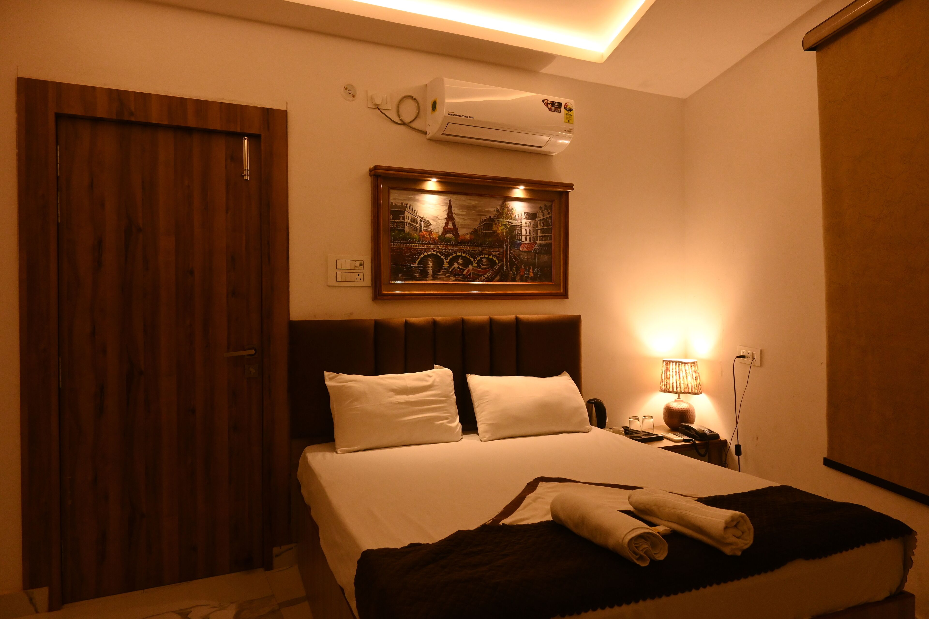 A hotel room with a bed, a nightstand, a lamp, an air conditioner, and a door.