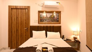 A hotel room with a wooden door, a bed with white bedding, a painting of a cityscape, a bedside table with a lamp, and a wall-mounted air conditioner. - Brij Inn (Varanasi)