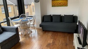 En moderne stue med en grå sofa, et lille spisebord med stole, et fjernsyn på et stativ og et billede af solsikker på væggen.