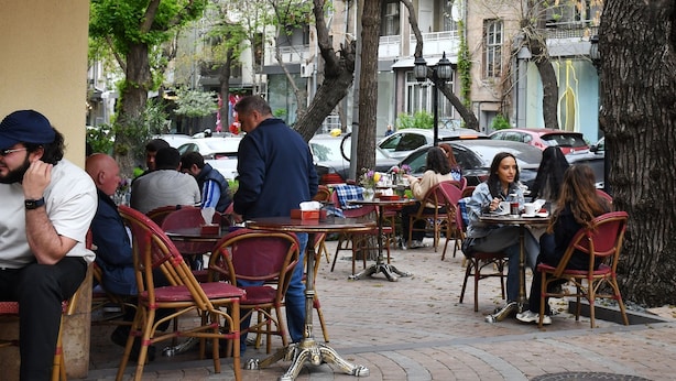 Um café ao ar livre com mesas e cadeiras, rodeado por árvores e edifícios.