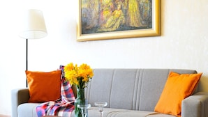Uma sala de estar com um sofá cinza, almofadas laranja, uma mesa de centro de madeira com um vaso de flores amarelas e um quadro na parede.