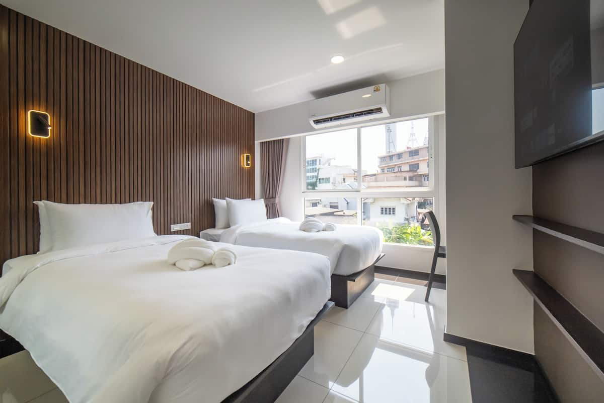 A hotel room with two beds, a large window with a view of buildings, an air conditioning unit, and a flat-screen TV mounted on the wall.