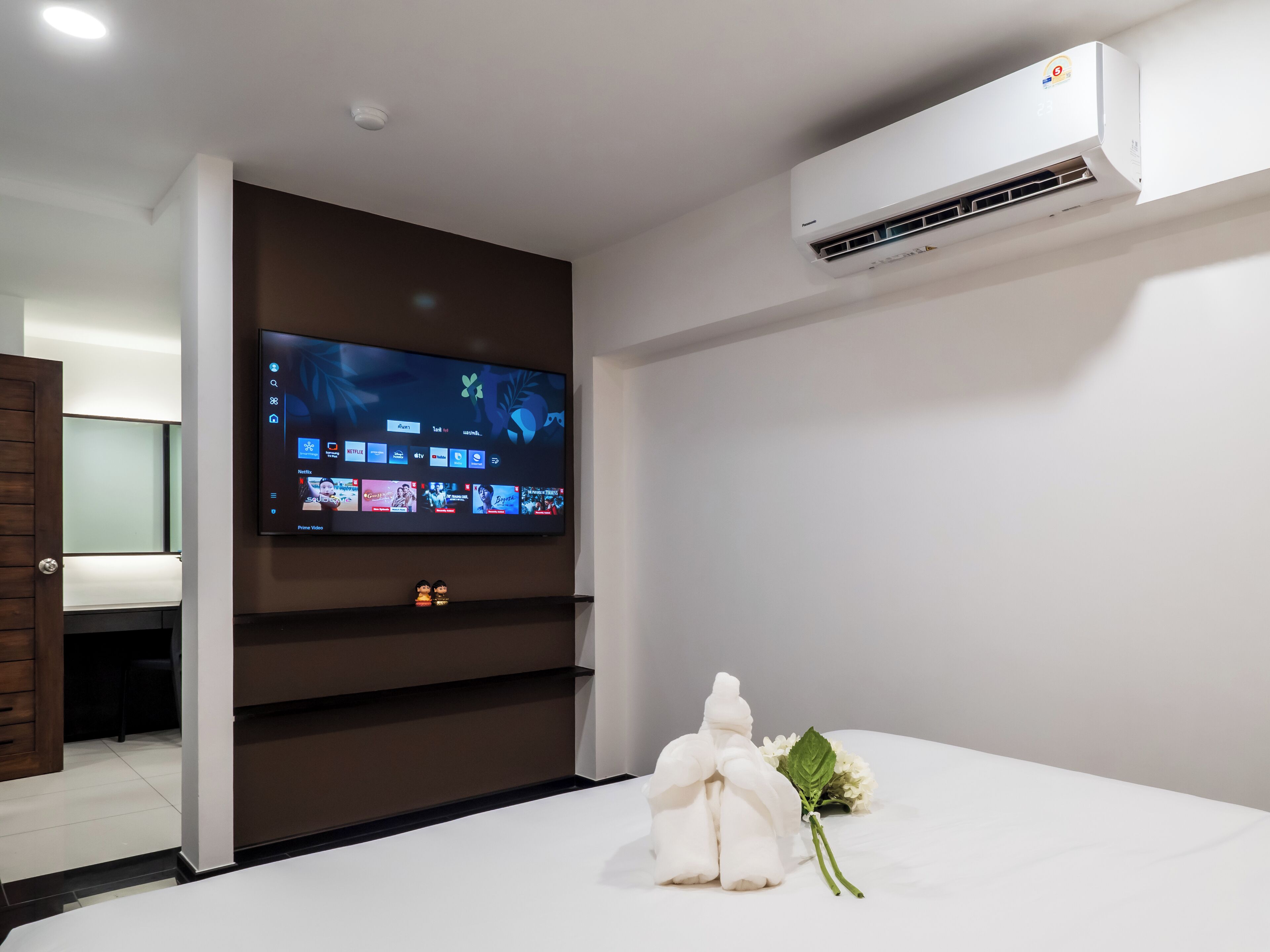 A hotel room with a flat-screen TV mounted on the wall, a built-in air conditioning unit, a wooden wardrobe, and a bed with a white towel and a flower on it.