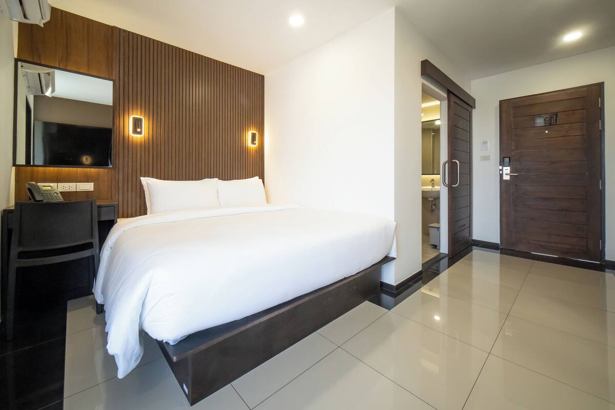 A modern hotel room with a large bed, a desk with a telephone, a mirror, and a wooden panel wall.