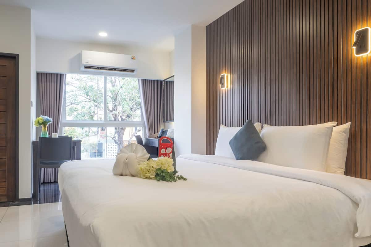 A hotel room with a large bed, a wooden panel wall, a bedside table with a phone and a red object, and a window with curtains.