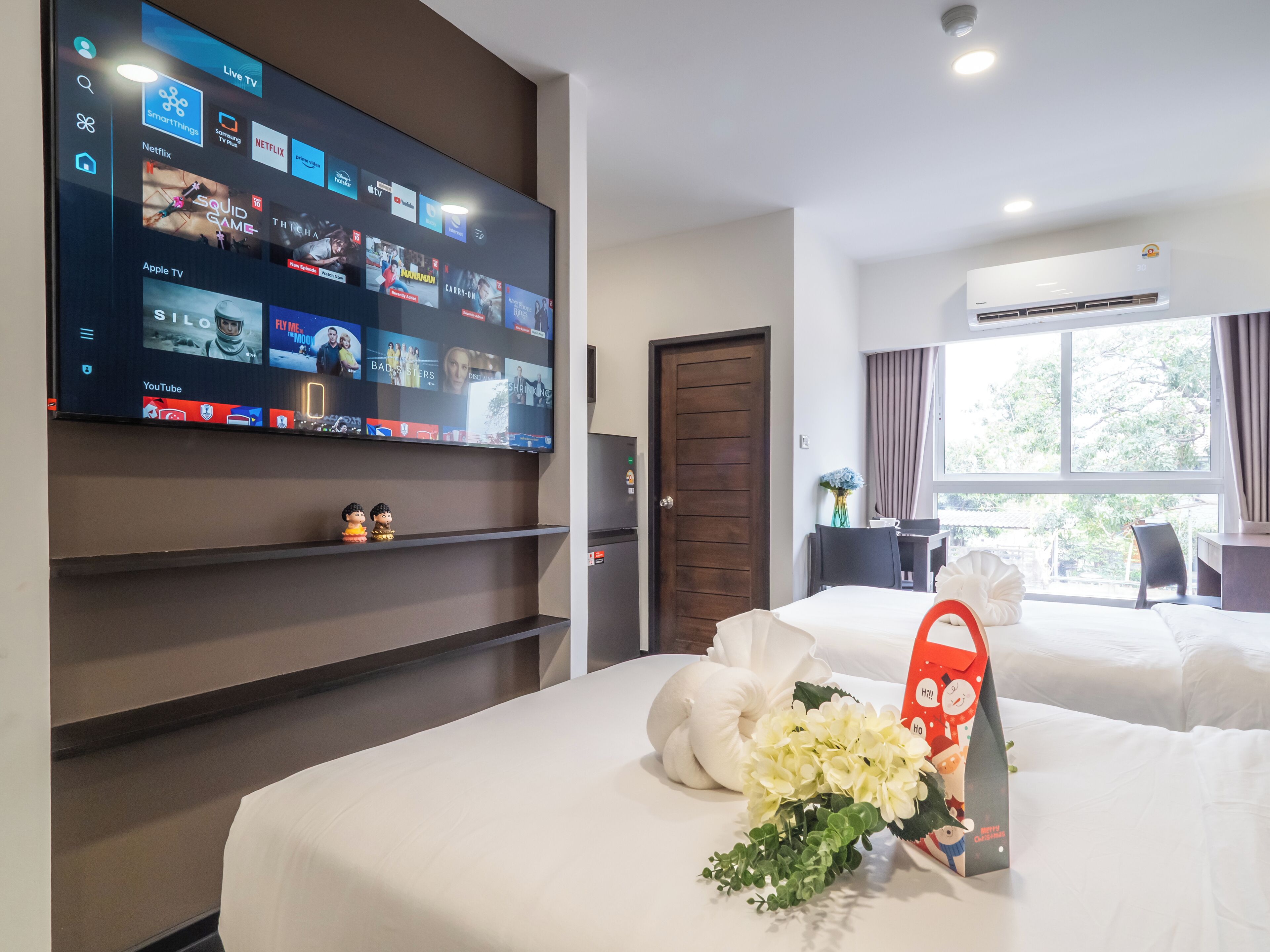 Una camera d'albergo con una grande TV che mostra diversi servizi di streaming, un letto con biancheria bianca, un tavolino con un vaso di fiori e una finestra con tende.