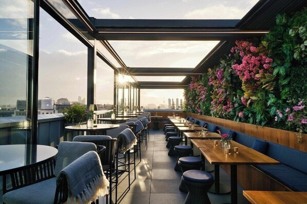 Een dakrestaurant met glazen overkapping, terras en een wand vol groen en roze bloemen.