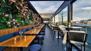 Ein Essbereich im Freien mit Holztischen und blau gepolsterten Sitzgelegenheiten, eine vertikale Begrünung mit rosa Blüten und ein Blick auf die Stadt durch große Fenster.