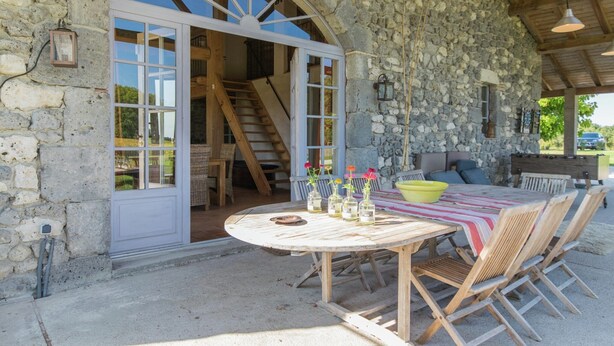 Un espace de restauration en plein air agrémenté d’un mur en pierre, de mobilier en bois et offrant une vue sur l’intérieur par le biais d’une porte en arc de cercle.