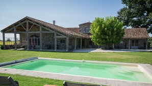 Un espace piscine avec des chaises longues et un patio couvert, attenant à un bâtiment en pierre surmonté d’un toit en tuiles.