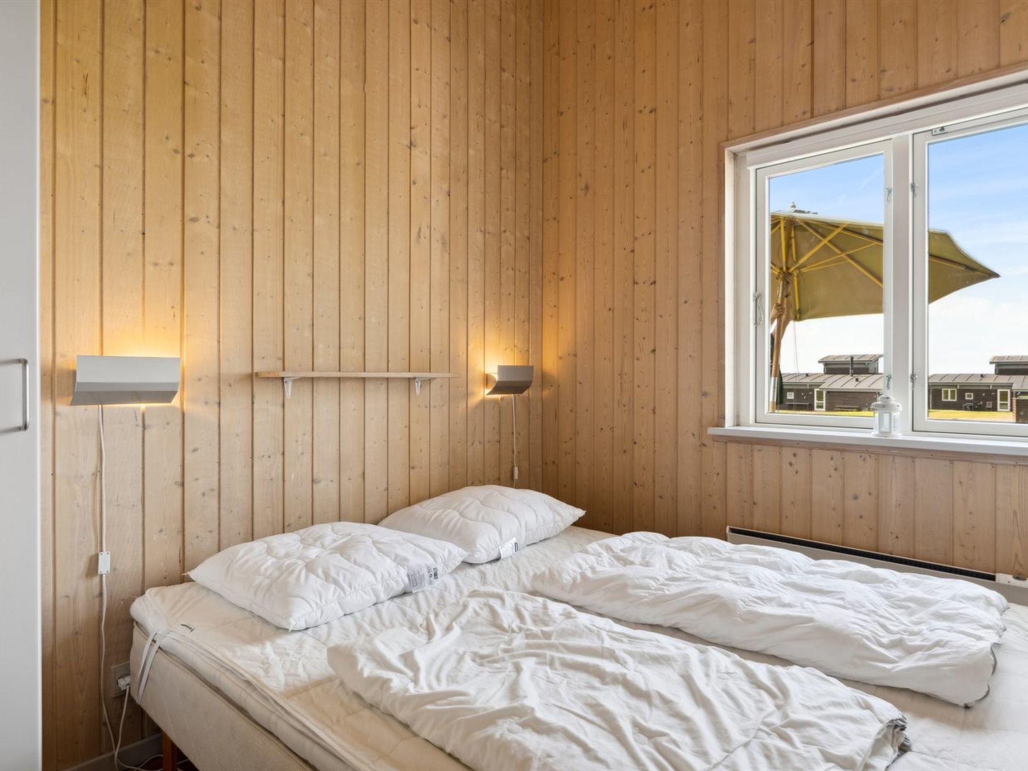 Camera da letto con pareti in legno, un letto con biancheria bianca, due lampade a parete e una finestra con un ombrello.