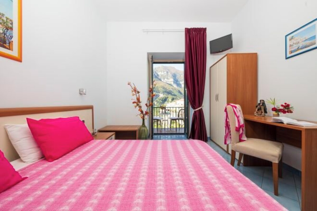 A hotel room with a bed, a desk, a chair, a wardrobe, and a balcony with a view of the mountains.