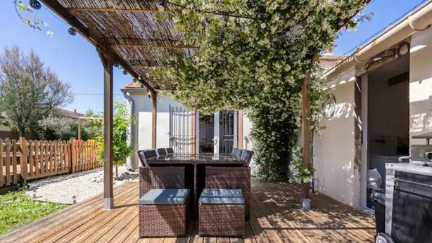 Un patio exterior con terraza de madera, una mesa con sillas y una pérgola que proporciona sombra.