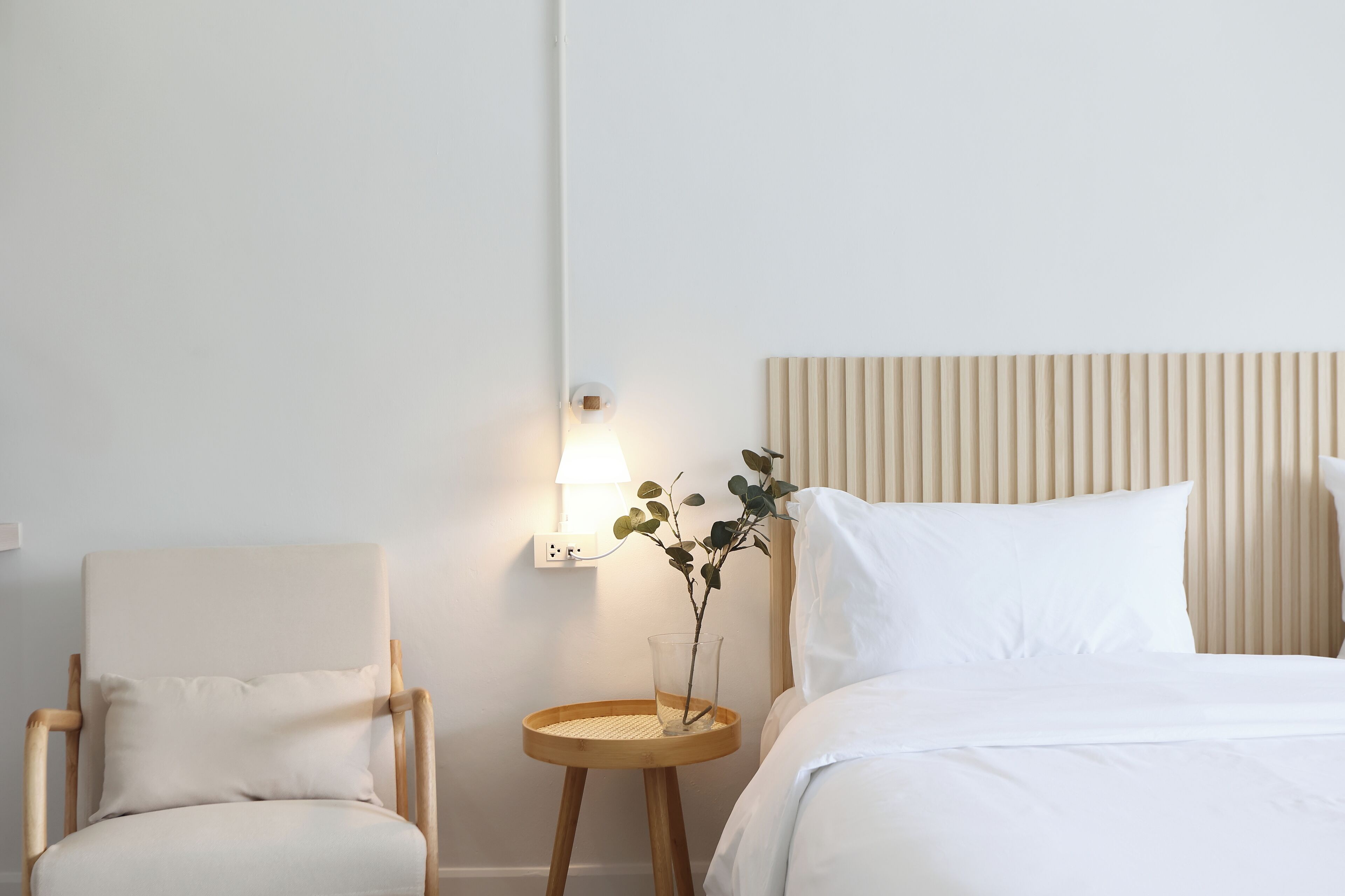 Una habitación de hotel con una cama, una silla, una mesita de noche con lámpara y un enchufe en la pared.