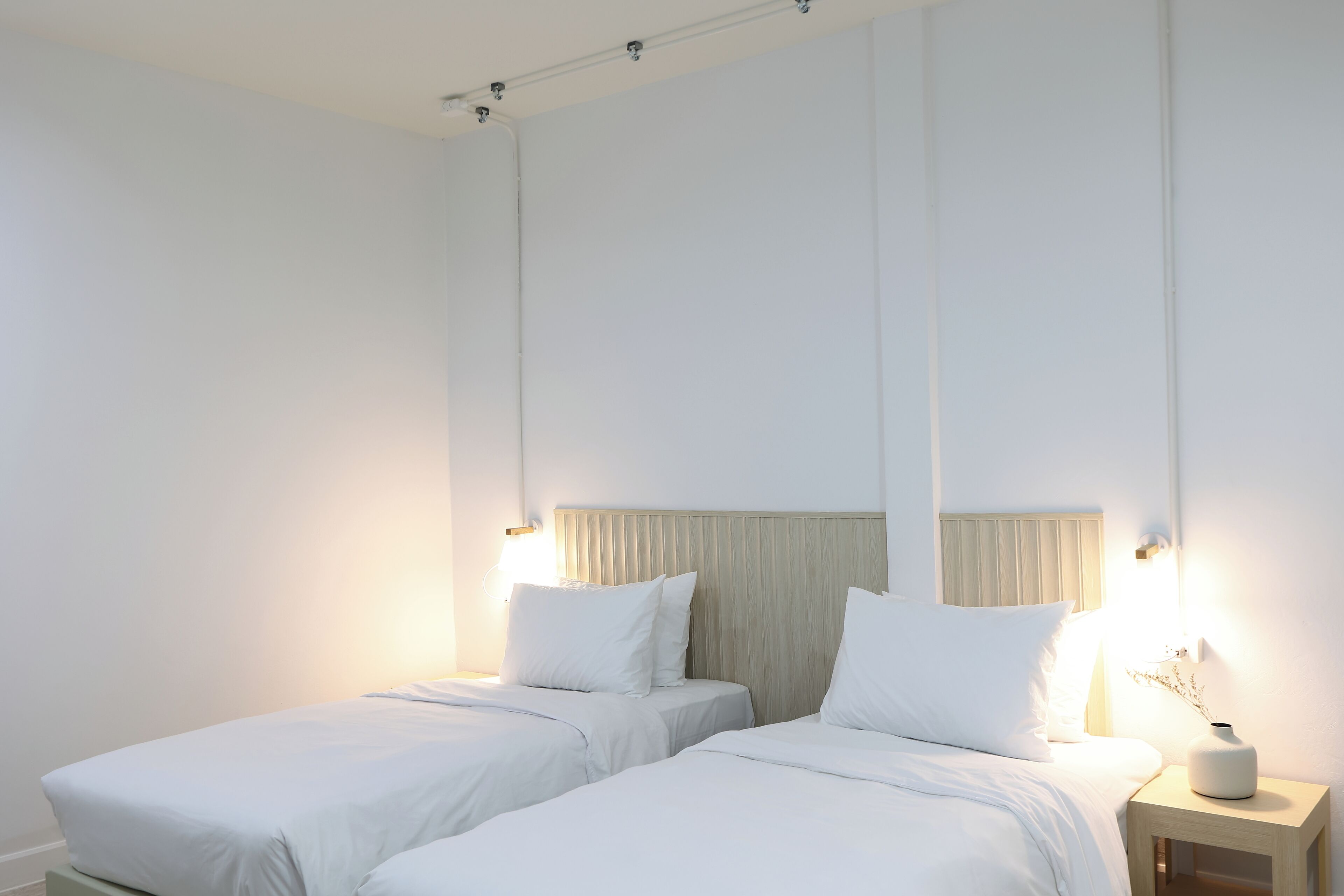 Et minimalistisk hotelværelse med to senge, et natbord med lampe, og en vægmonteret lysarmatur.