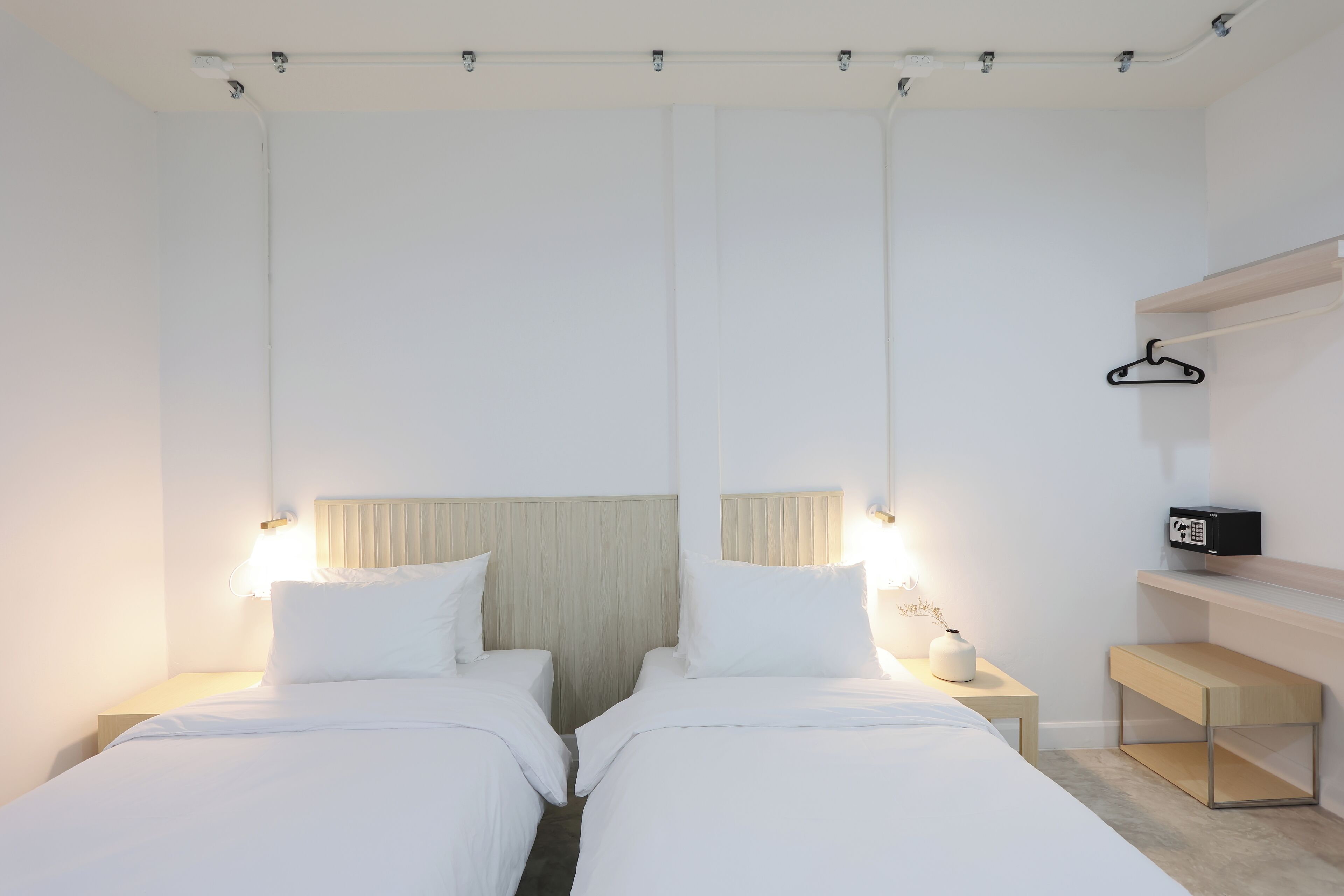 Une chambre d’hôtel moderne avec deux lits simples, une table de chevet avec une lampe, une petite étagère avec une horloge et un porte-manteau fixé au mur.
