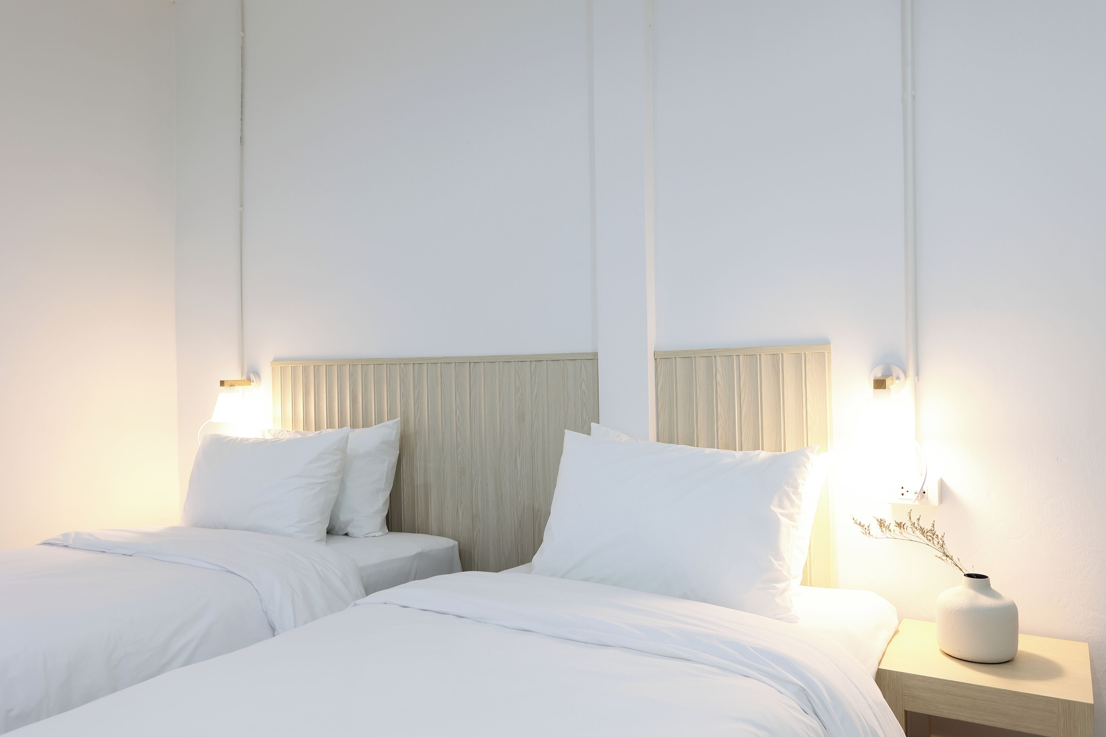A hotel room with two beds, a bedside table with a vase, and a wall-mounted light fixture.