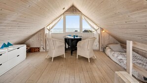 Een gezellig zolderkamertje met een houten schuine plafond, een klein tafeltje met stoelen, een raam met uitzicht op de zee, en een bed met een bloemenbedekking.