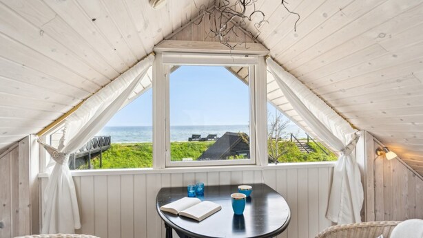 Een gezellige kamer met een houten plafond, een tafel met stoelen en een groot raam met uitzicht op een grasveld en de zee.