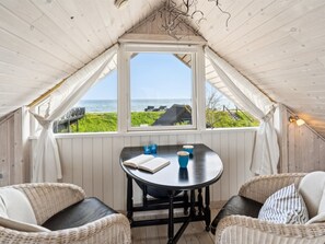 Ein gemütliches Zimmer mit Holzbalkendecke, einem Tisch mit Stühlen und einem großen Fenster mit Blick auf eine Wiese und das Meer.