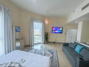 A modern hotel room with a bed, a sofa, a TV mounted on the wall, a large window with curtains, and a potted plant. - Snug Studio With City Views, Perfect For Couples (Dubai)