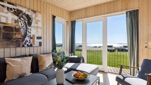 A modern living room with a sofa, a coffee table with fruit, and a view of a green field and water.