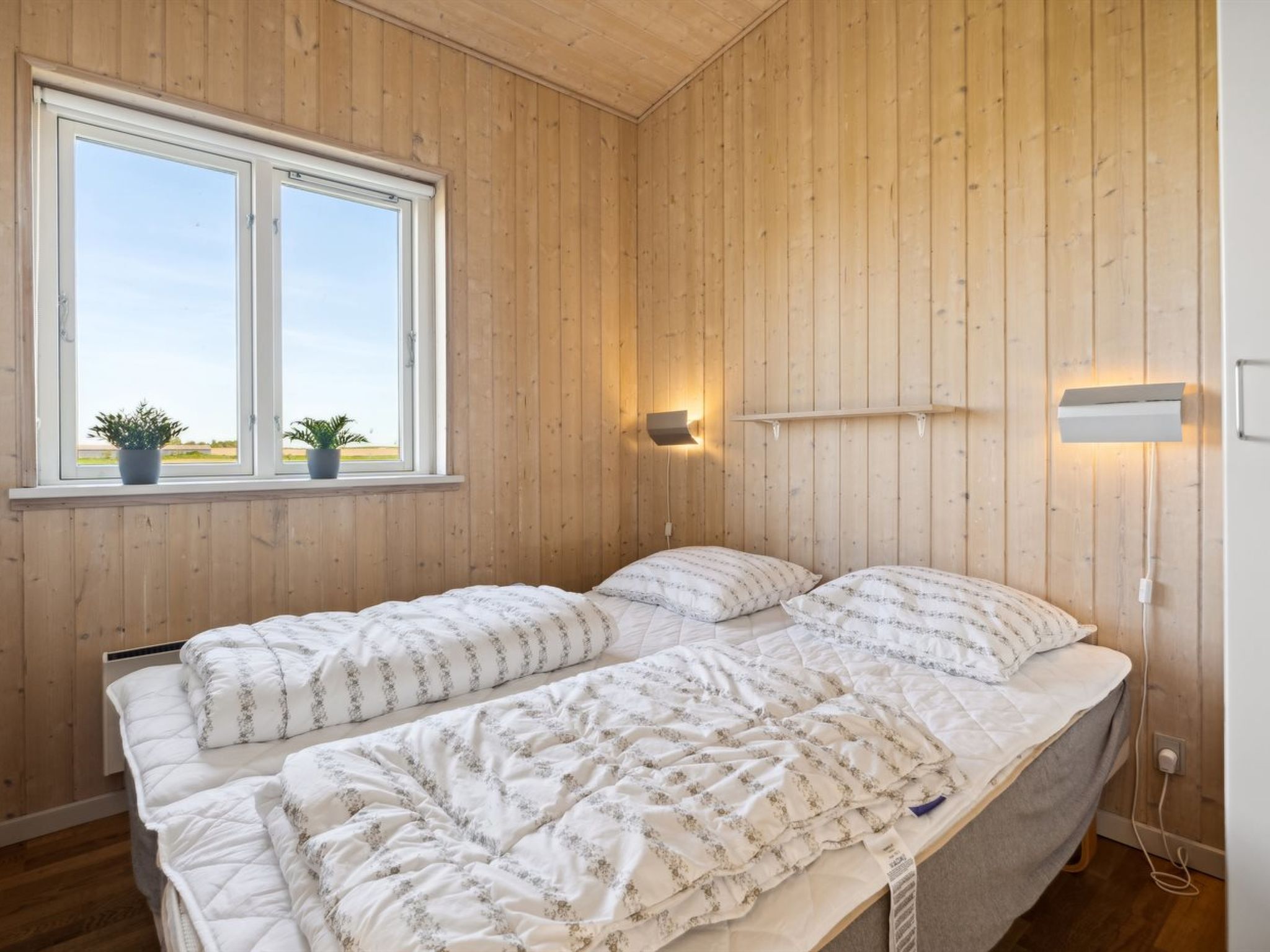 Une chambre avec des murs en bois, une grande fenêtre agrémentée de deux plantes en pot, un lit recouvert d’une couette blanche et deux appliques murales.