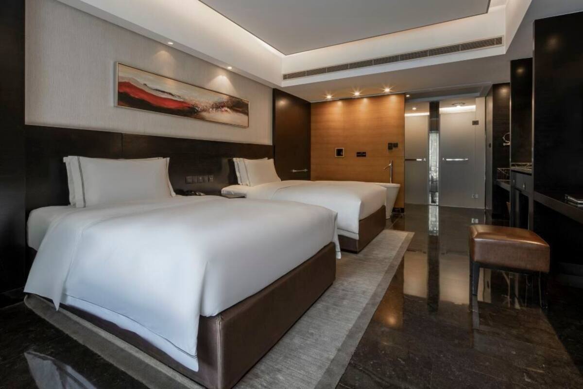 Ein modernes Hotelzimmer mit zwei Betten, einem großen Gemälde, einem Flachbildfernseher und einem durch eine offene Tür einsehbaren Badezimmer.