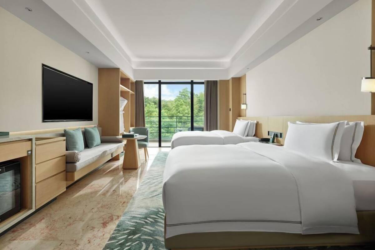 Ein modernes Hotelzimmer mit einem großen Bett, einer Couch, einem Fernseher und einem Balkon mit Blick auf Bäume.