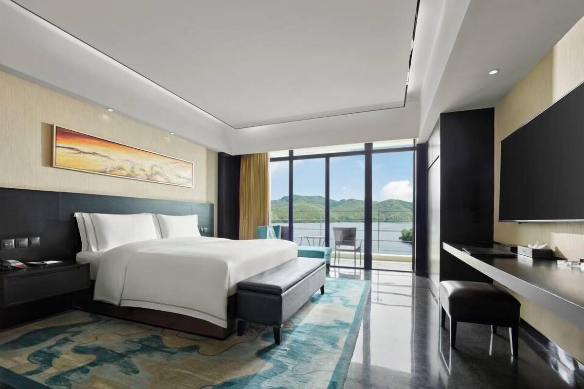 Ein modernes Hotelzimmer mit einem großen Bett, einem Schreibtisch, einem Sessel und einem Fernseher. An der Wand hängt ein Gemälde und es gibt einen Blick ins Freie.