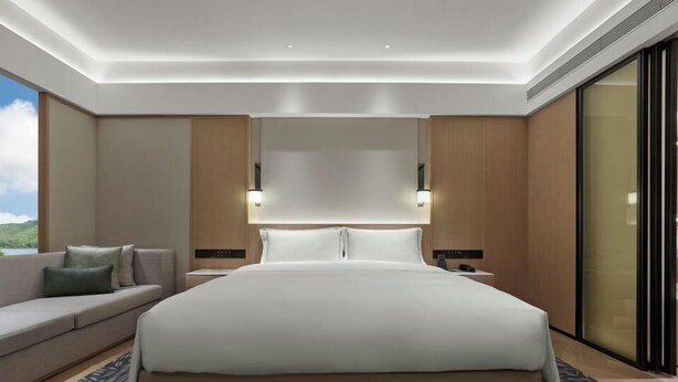 Quarto de hotel moderno com uma cama grande, um sofá, uma mesa de cabeceira e vista para o exterior.