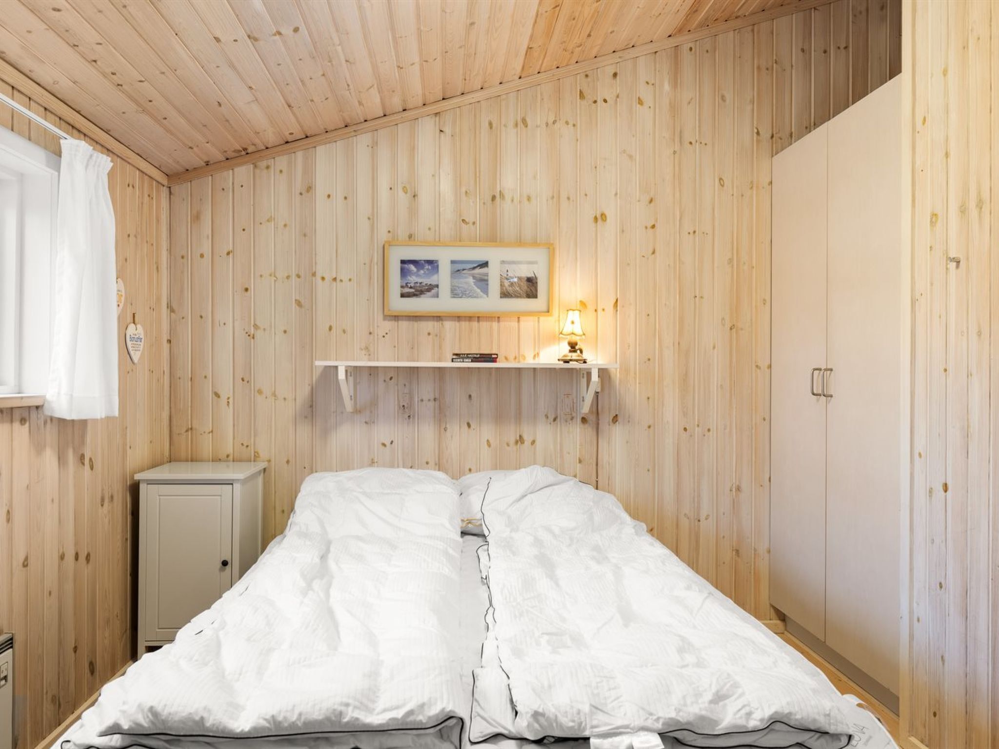 Ein Zimmer mit Holzwandverkleidung, einem Bett mit weißer Bettwäsche, einer kleinen Kommode und einem Regal mit Lampe und Bildern.