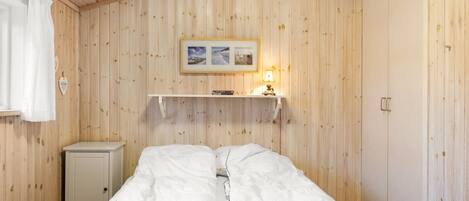 Ein Zimmer mit Holzwandverkleidung, einem Bett mit weißer Bettwäsche, einer kleinen Kommode und einem Regal mit Lampe und Bildern.