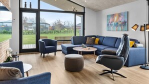 Ein modernes Wohnzimmer mit einem großen Glastür-Zugang zu einem Garten, einem blauen Sofagarnitur, einem Ledersessel und einem Gemälde an der Wand.