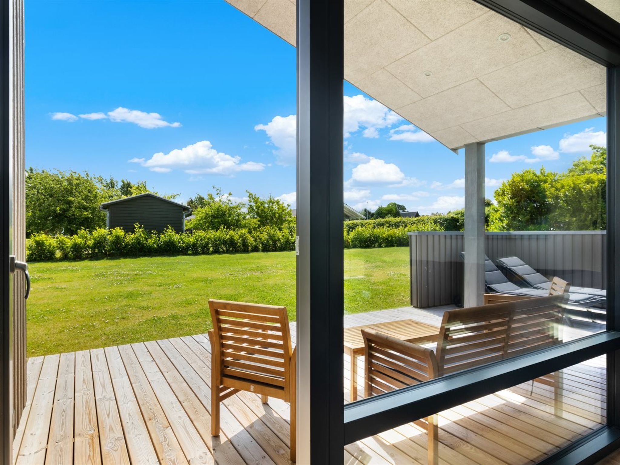 Una terraza moderna al aire libre con mobiliario de madera y una vista despejada de un césped verde y un cobertizo pequeño al fondo.