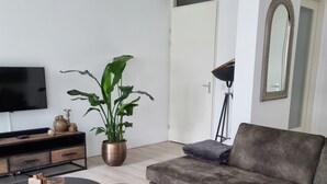 Een moderne woonkamer met een grijze bank, twee ronde zwarte tafels, een grote potplant, een flatscreen televisie en een vloerlamp.