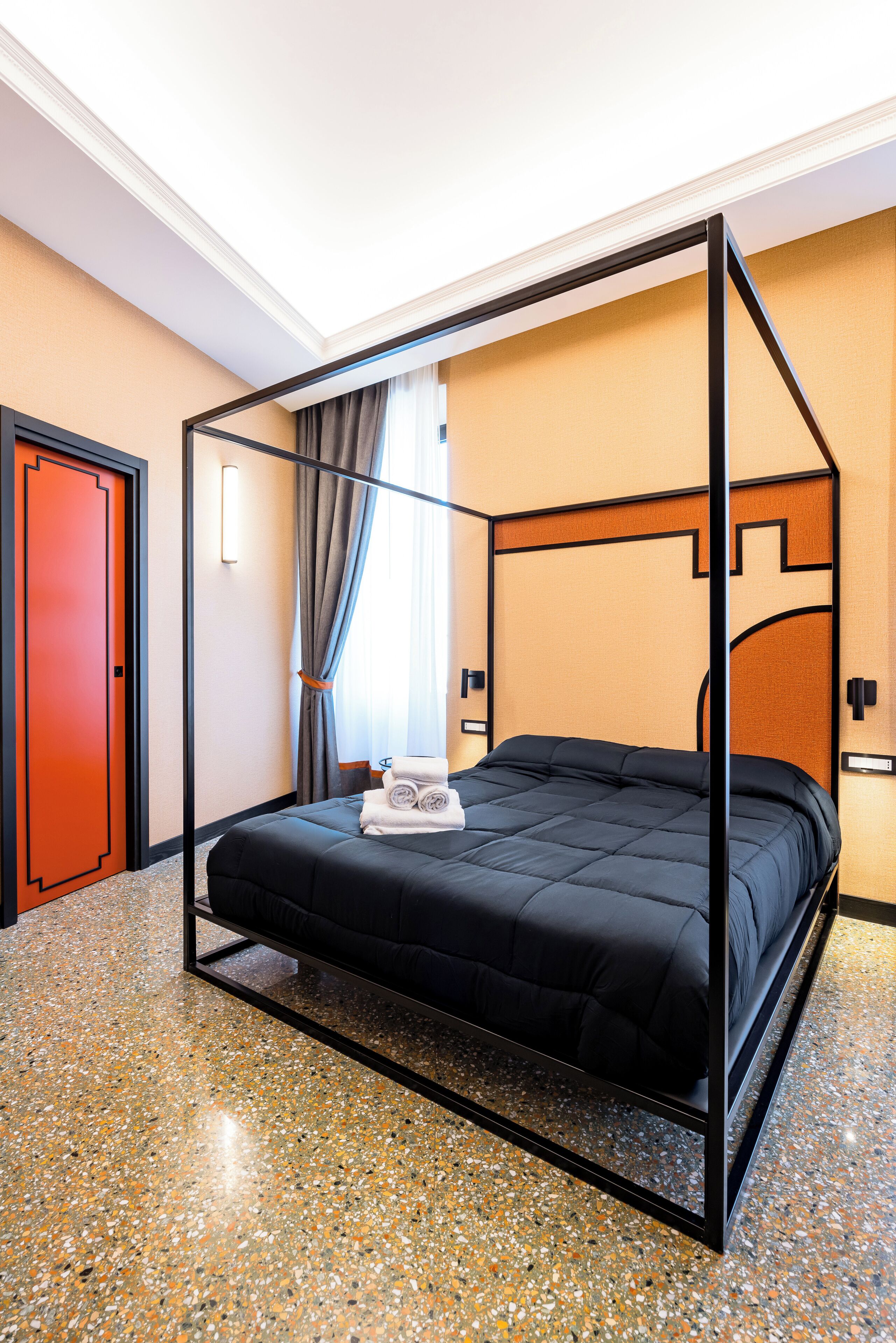 A four-poster bed with a black frame and a dark blue bedspread, a red door, and a mirror on the wall.