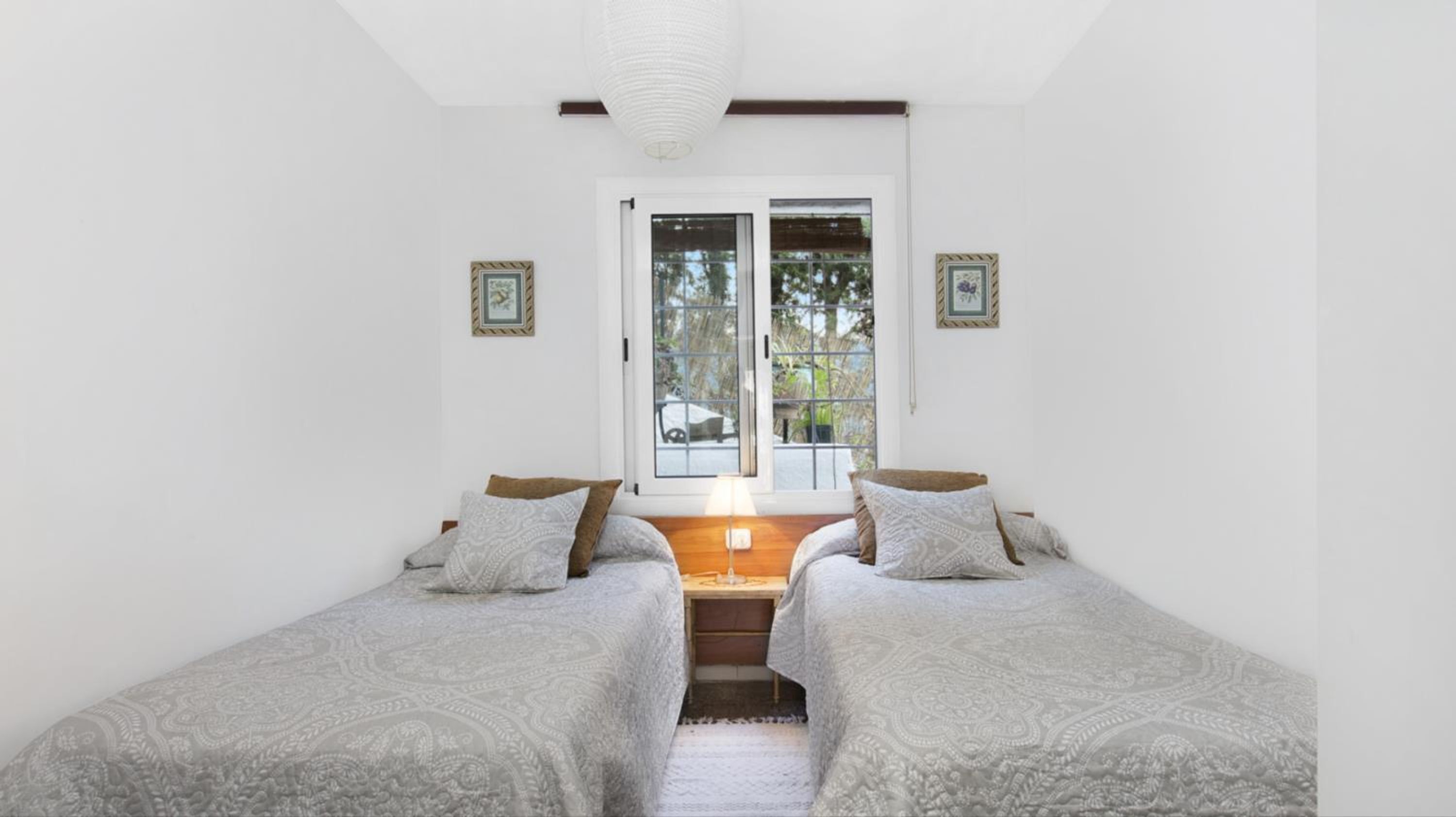 A bedroom with two single beds, a window with a view of greenery, and a small desk with a lamp.