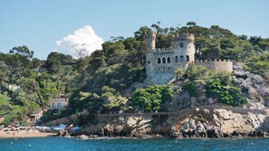 A coastal castle with a stone tower, surrounded by lush greenery and a beach with boats. - Palmeras in Lloret de Mar (Lloret de Mar)