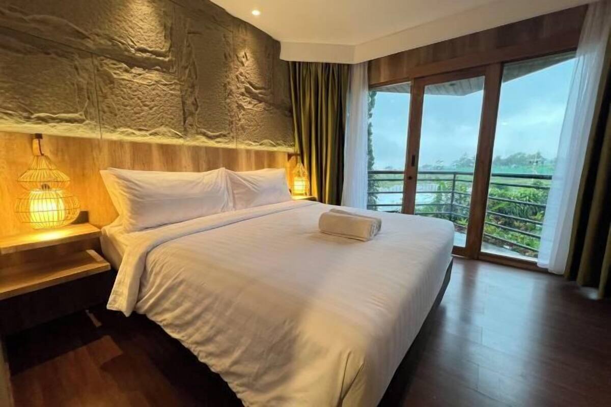 A hotel room with a large bed, wooden headboard, and a view of greenery through the windows.