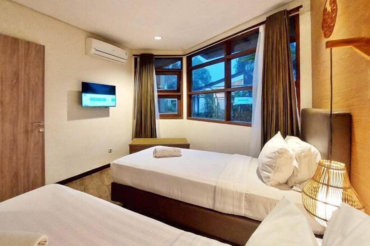 A hotel room with two beds, a TV, a window with curtains, and an air conditioning unit.