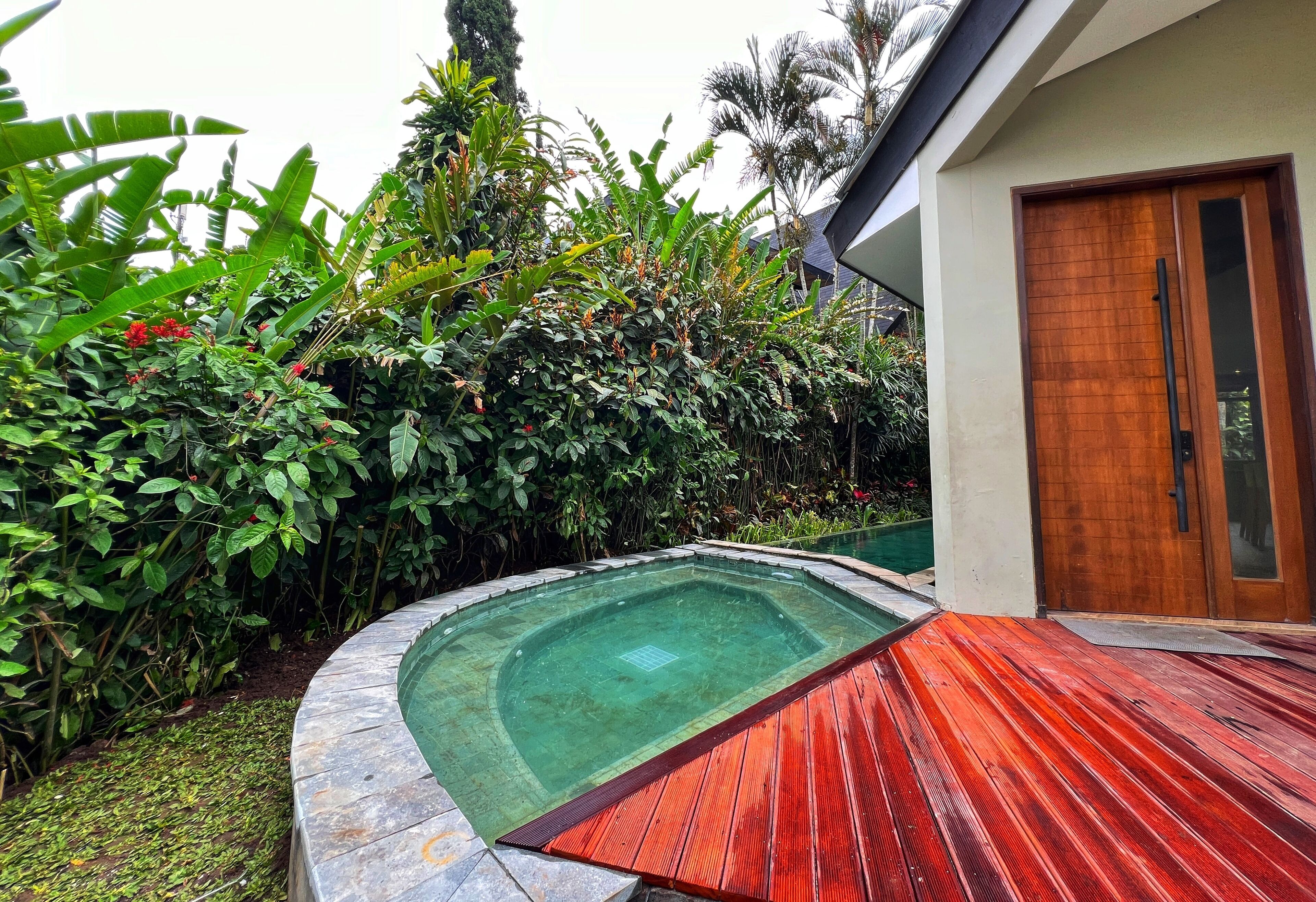Una piscina con cubierta de madera, rodeada de exuberante vegetación y un edificio moderno con puerta de madera.