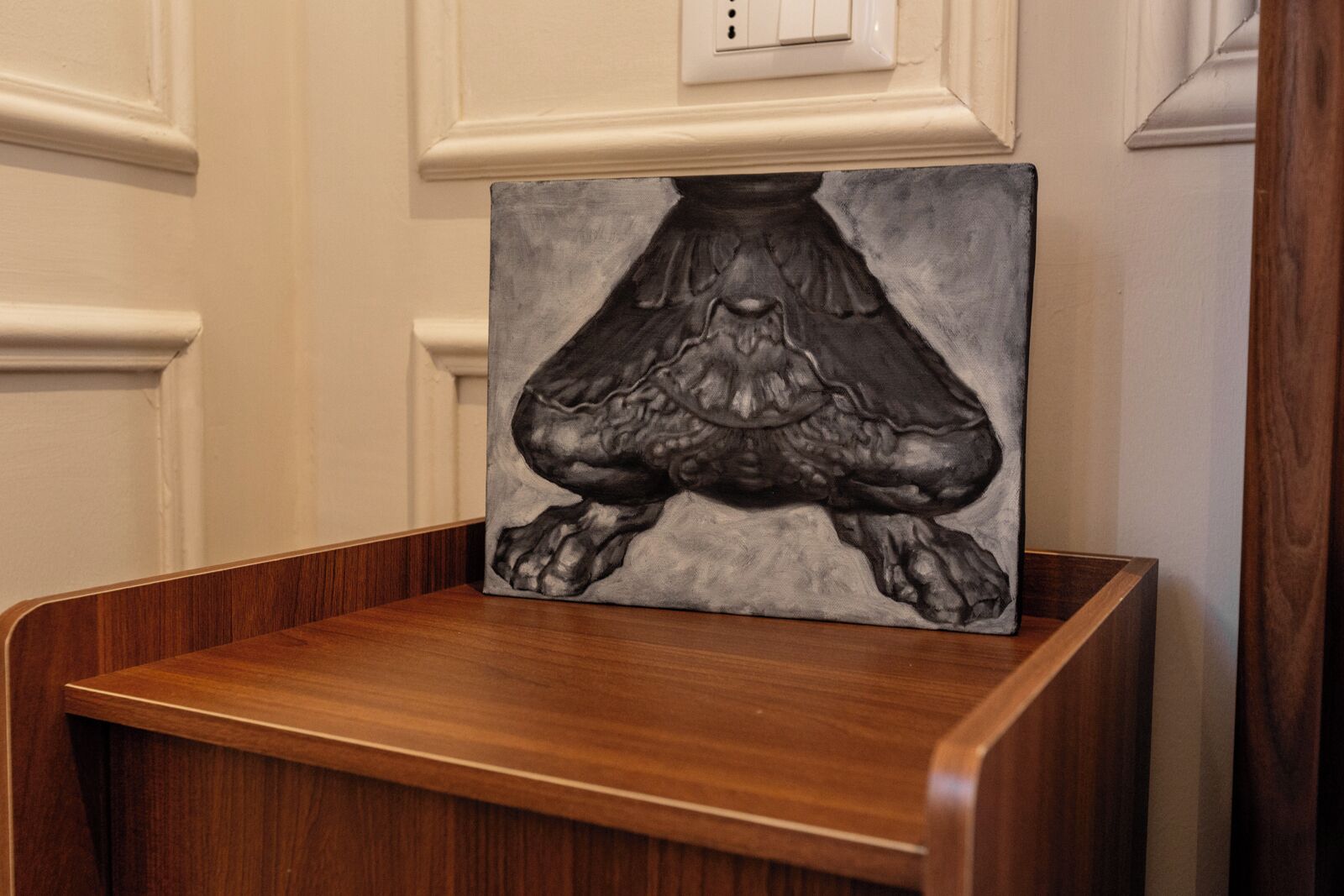 Een ingelijste afbeelding van een menselijke figuur op een houten voetstuk, voor een muur met een lichtschakelaar.