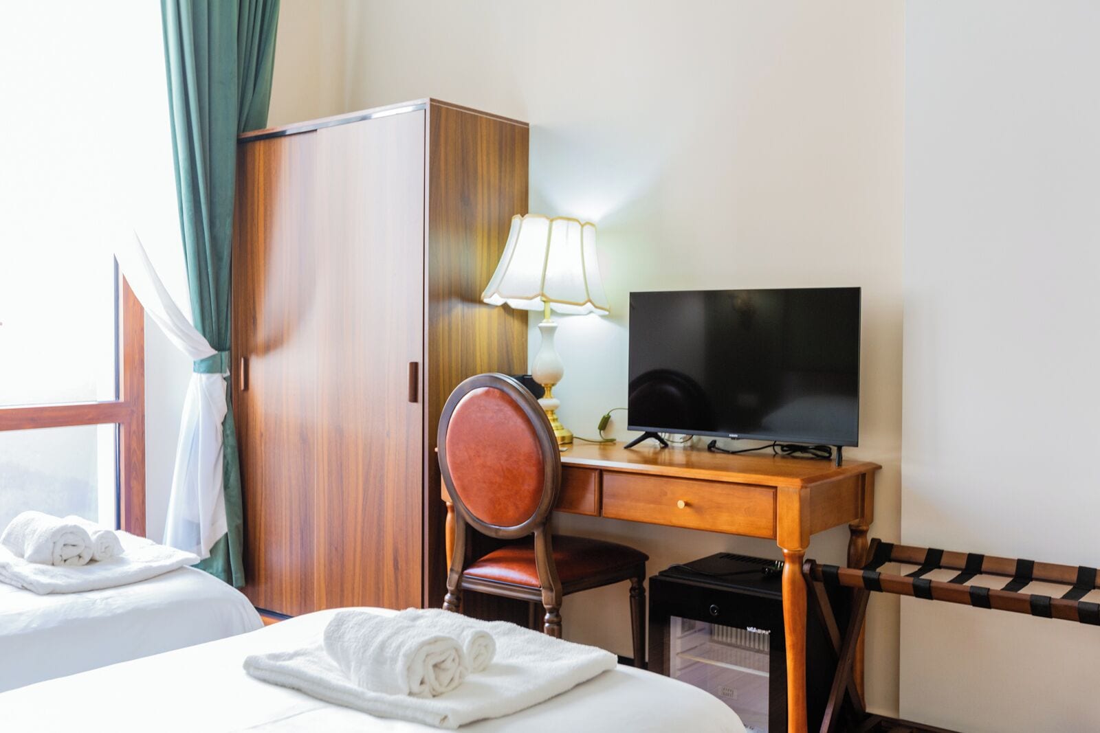 A hotel room with two beds, a wooden desk with a television, a chair, a lamp, and a minibar.