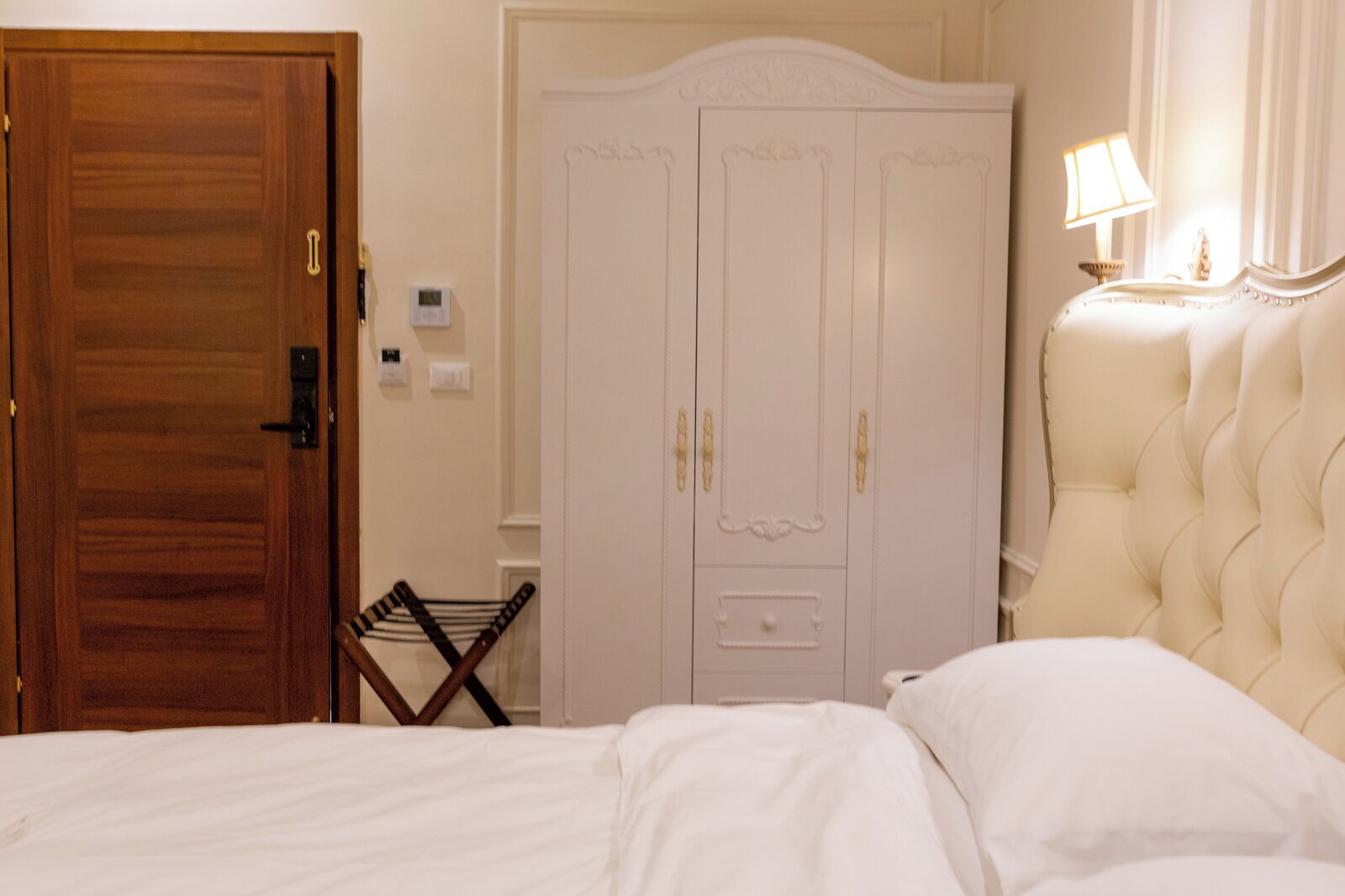 A bedroom with a wooden door, a white wardrobe, a bed with white bedding, and a lamp on a bedside table.