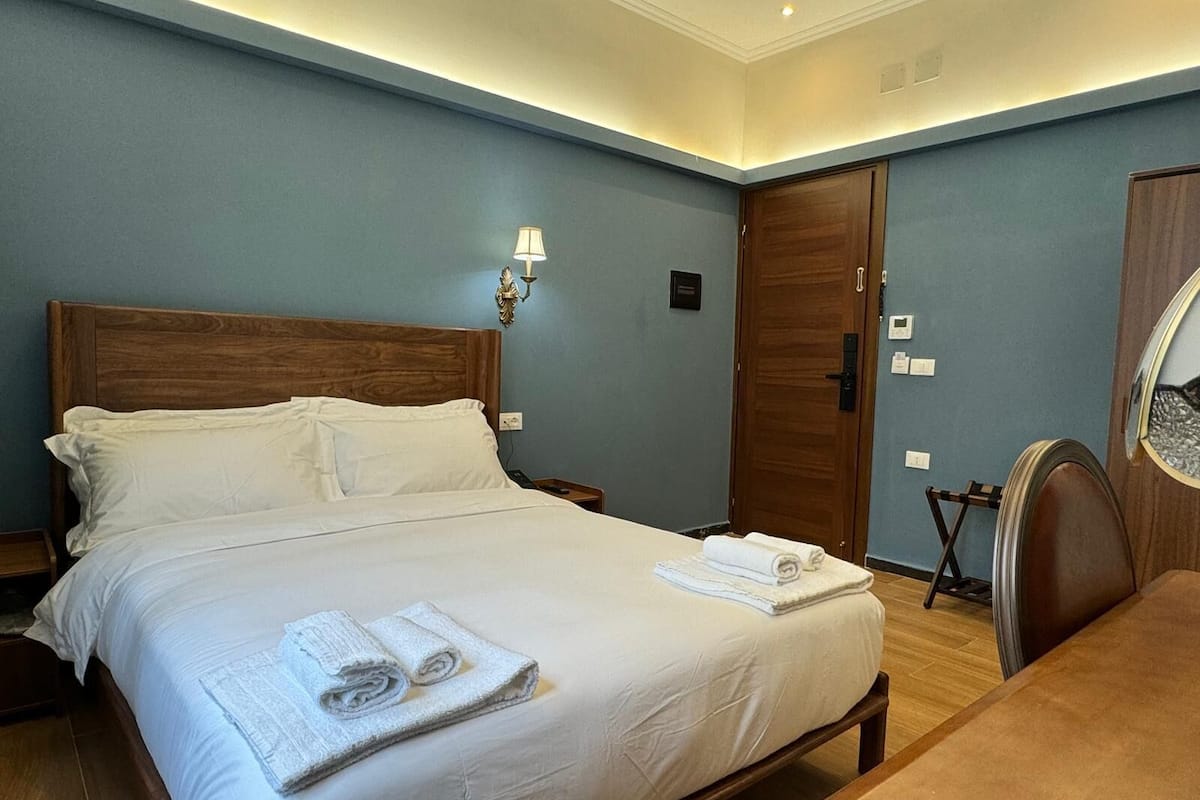 Una cama bien hecha con sábanas y toallas blancas, un cabecero de madera, una mesita de noche y una lámpara de pared.