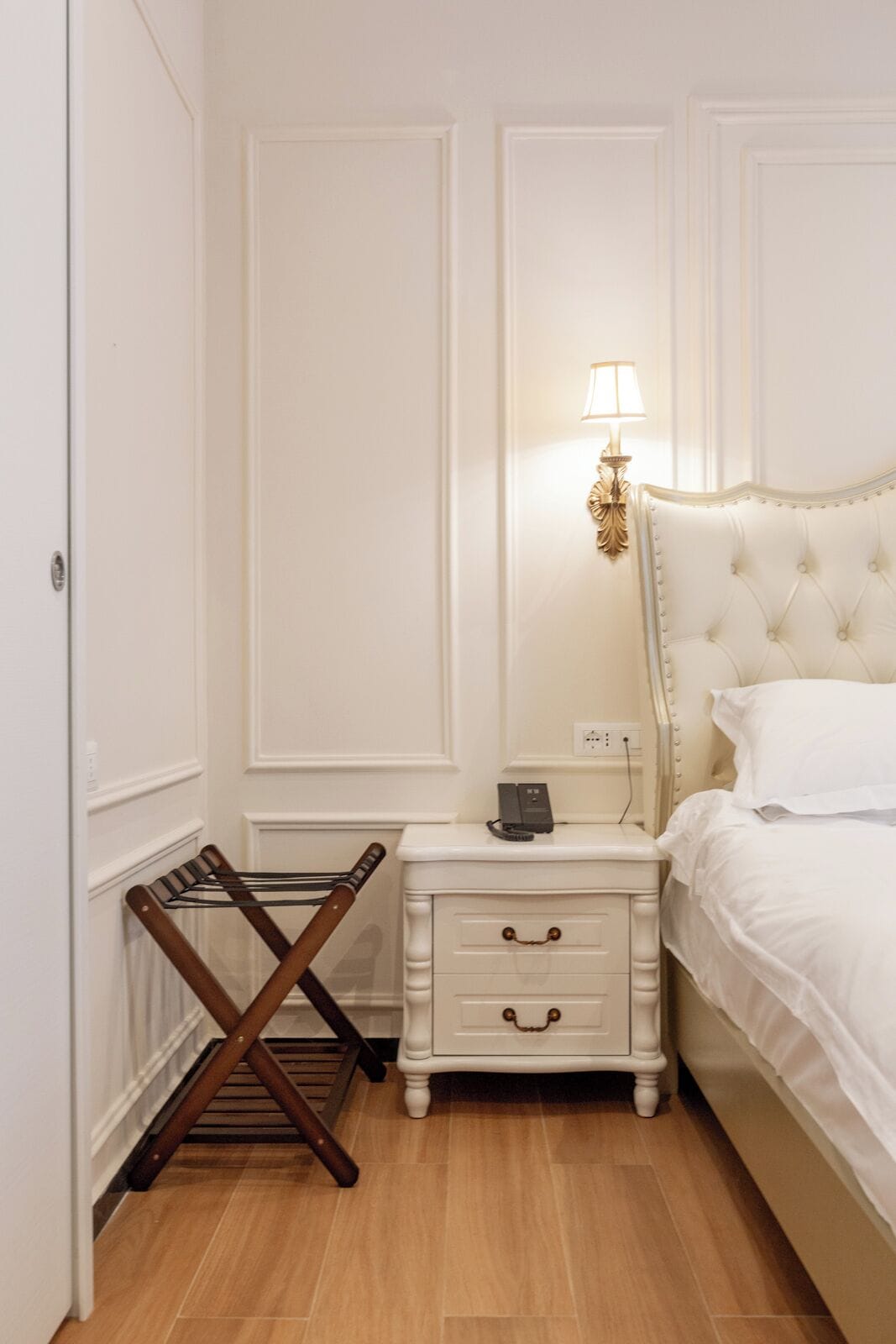 A neatly arranged bedroom with a bed, a bedside table with a lamp, a small drawer, and a wooden drying rack.